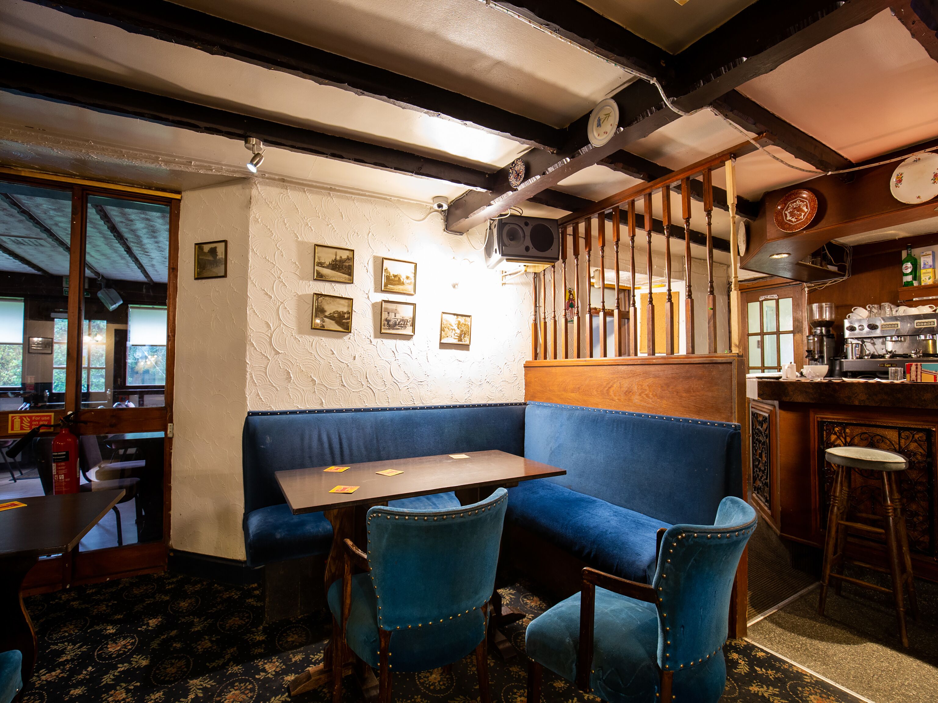 A cozy corner with a wooden table and blue upholstered chairs, a bar area with a coffee machine, and framed pictures on the wall.