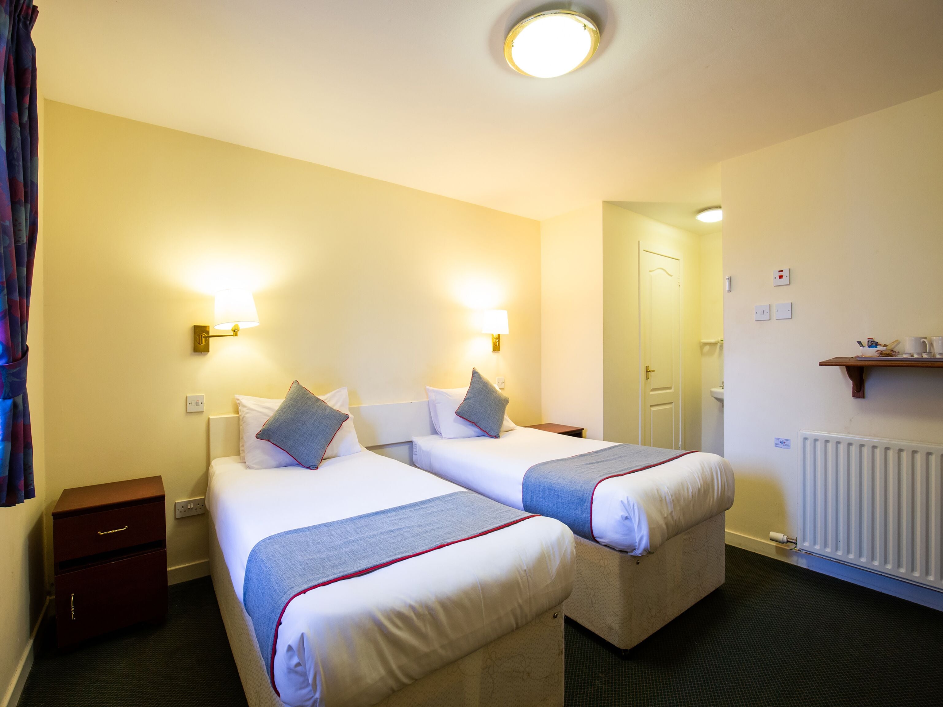 Quarto de hotel com duas camas, luminárias de cabeceira, mesa de apoio, radiador e uma porta que dá para outro cômodo.