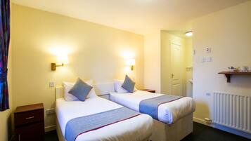 Quarto de hotel com duas camas, luminárias de cabeceira, mesa de apoio, radiador e uma porta que dá para outro cômodo.