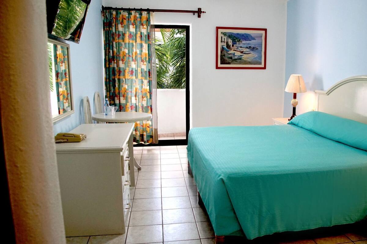 Un dormitorio con cama, escritorio, silla, ventana con cortinas, un cuadro en la pared y una lámpara.