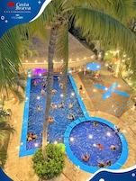 Un área de piscina con varias piscinas, palmeras y una estructura con techo de paja.