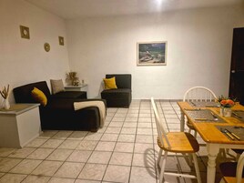 Una sala de estar con un sofá, una mesa de comedor con sillas, un cuadro en la pared y una mesita auxiliar con un jarrón.