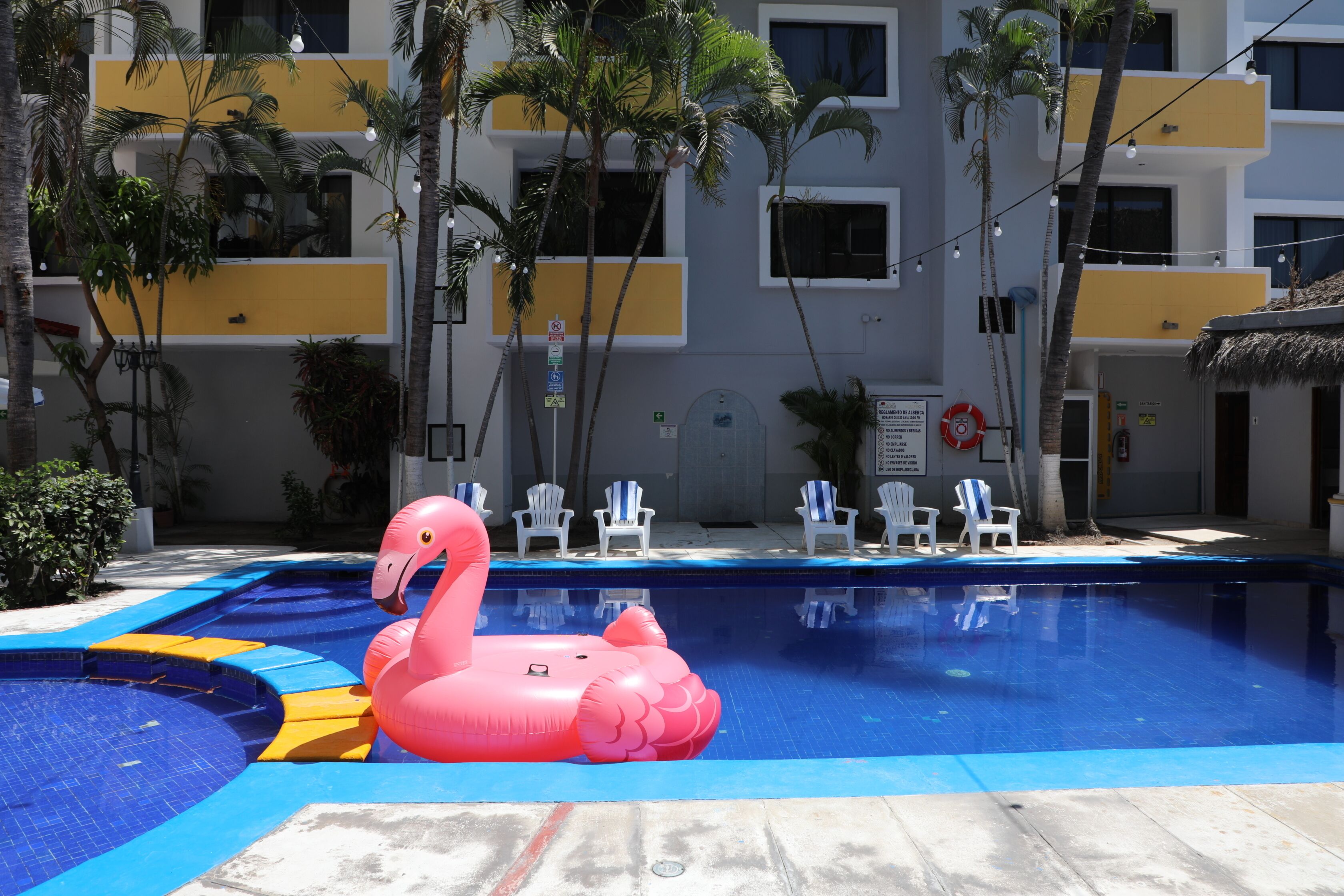 Una piscina con un flotador de flamenco rosa, rodeada de sillas blancas y palmeras.