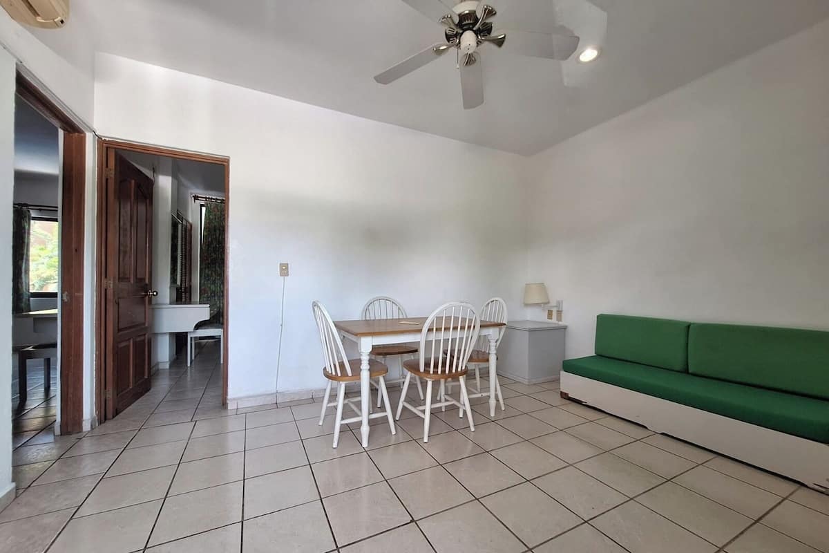 Una habitación con una mesa de comedor y sillas, un ventilador de techo, un sofá verde y suelo de baldosas.