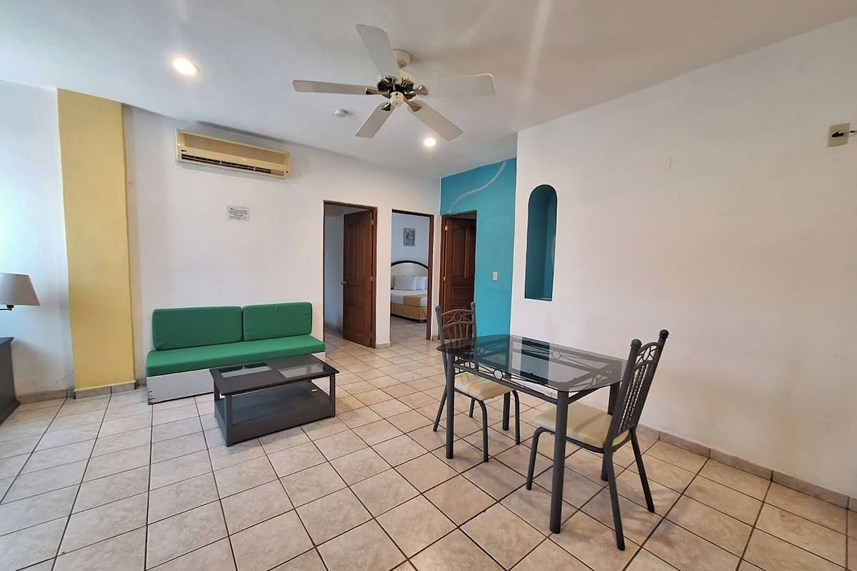 Una habitación con un sofá verde, una mesa de vidrio con sillas, un ventilador de techo y un equipo de aire acondicionado.