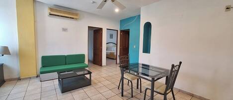 A room with a green sofa, a glass table with chairs, a ceiling fan, and an air conditioning unit.