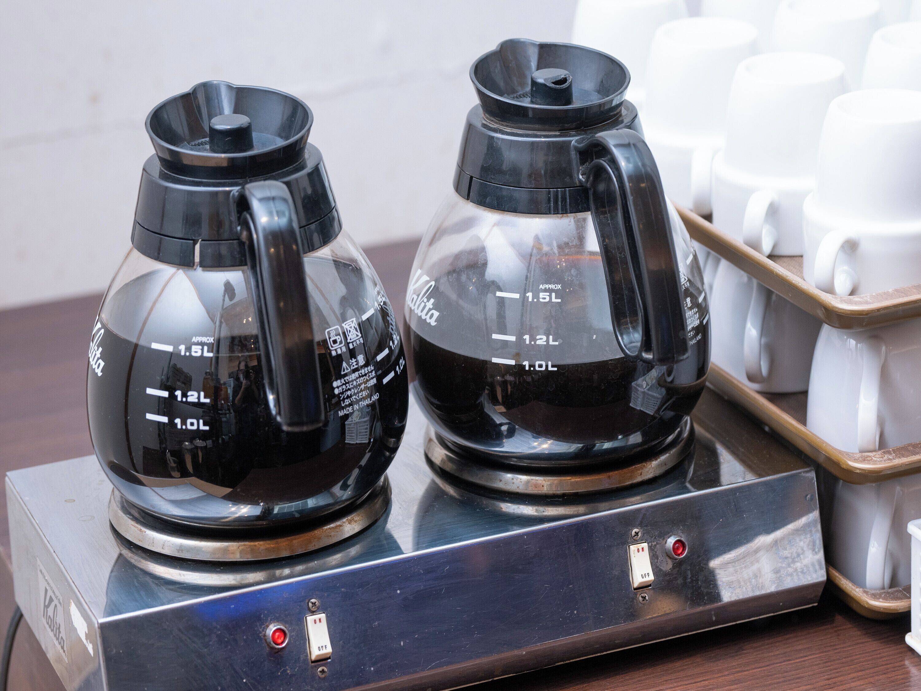 Two coffee makers on a metallic stand with measurement markings, a tray of white cups, and a switch.