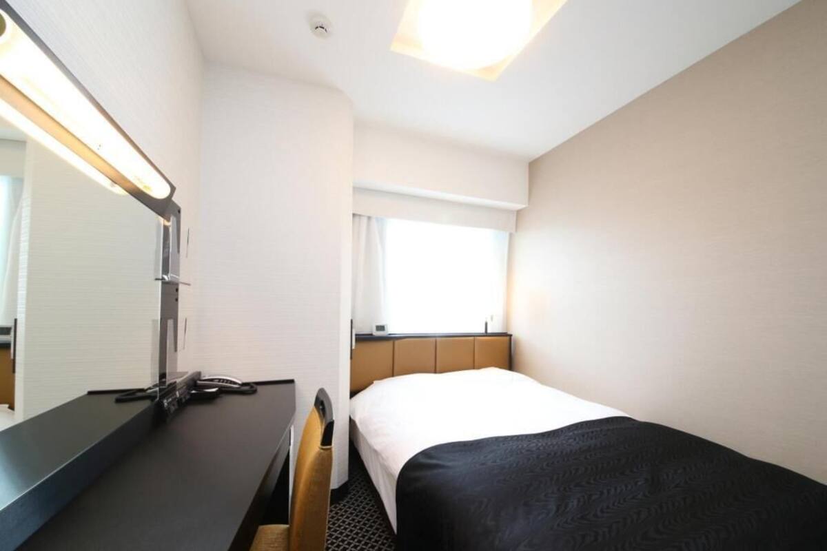 Una habitación de hotel con una cama, un escritorio con espejo, una ventana y una lámpara en el techo.