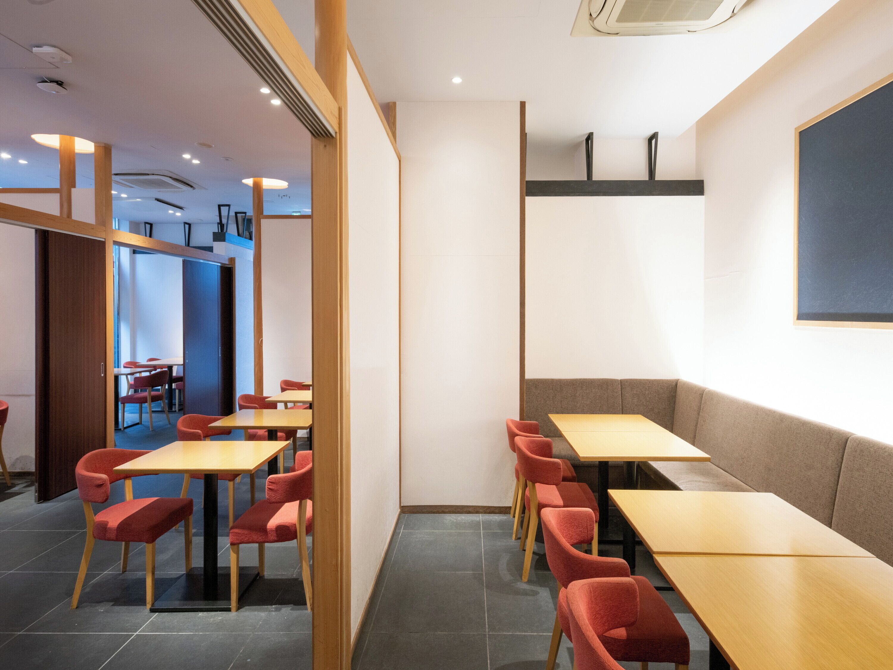 Ein modernes Restaurant mit Holztischen und roten Stühlen, eine große Kreidetafel an der Wand und Blick in einen weiteren Raum.