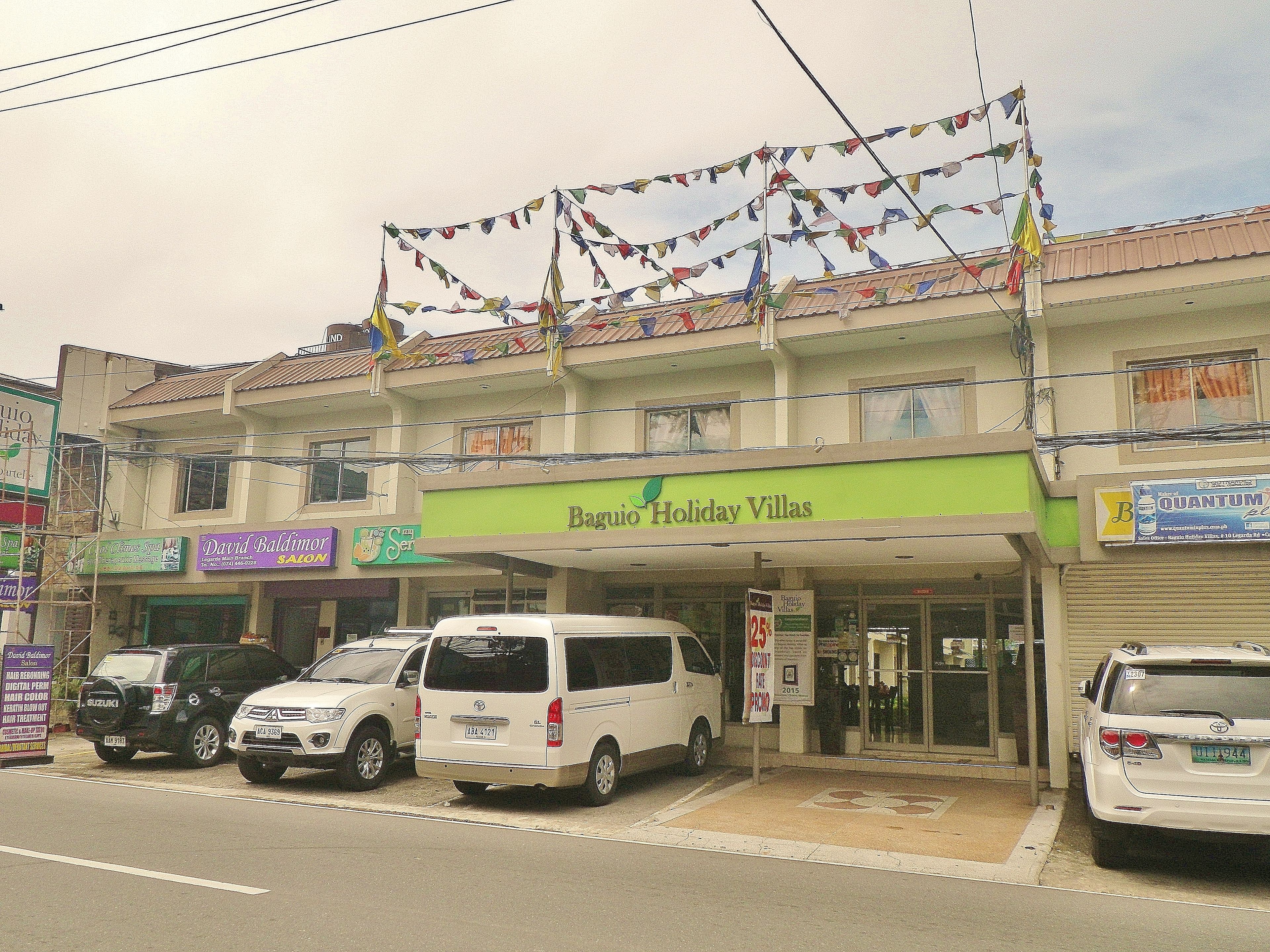 A building with a green sign reading "Baguio Holiday Villas". There are several cars parked in front, and a sign advertising a 25% discount.