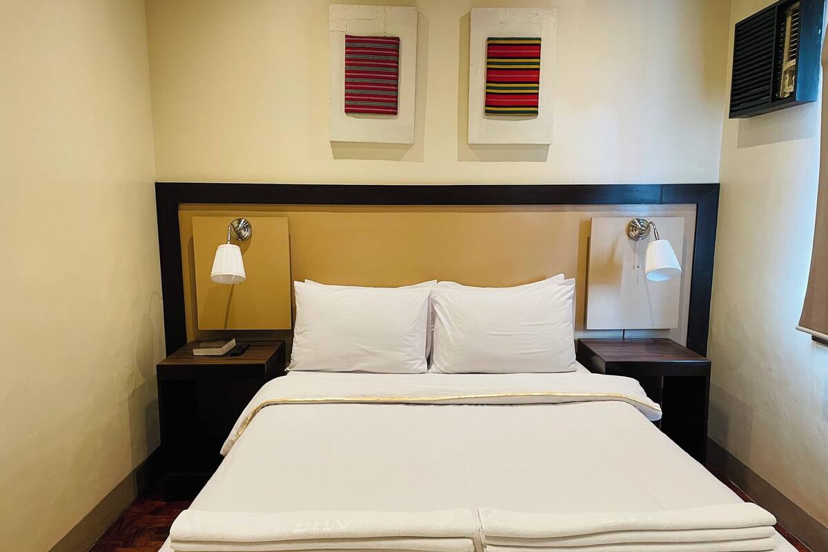 Een hotelkamer met een opgemaakt bed, twee nachtkastjes met lampen en twee wandventilatoren.