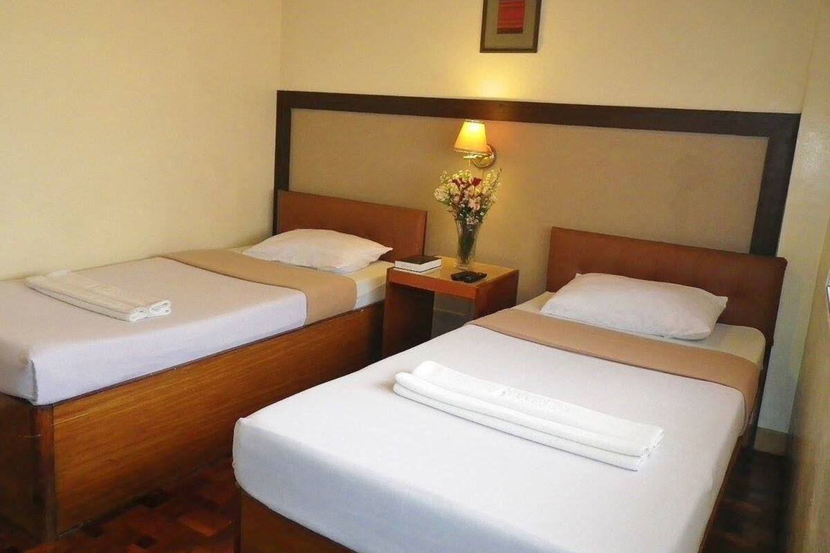 Een hotelkamer met twee enkele bedden, een nachtkastje met een lamp en bloemen, en een ingelijst schilderij aan de muur.
