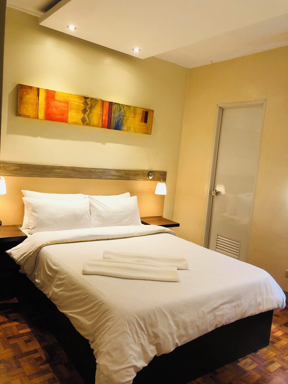 Habitación de hotel con una cama grande, dos lámparas de noche, un cabecero de madera, un cuadro abstracto colorido en la pared y una puerta con rejilla.
