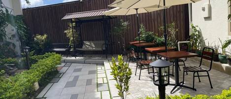 Un área de patio al aire libre con suelo de baldosas, mesas y sillas de madera, plantas en macetas y sombrillas que dan sombra.