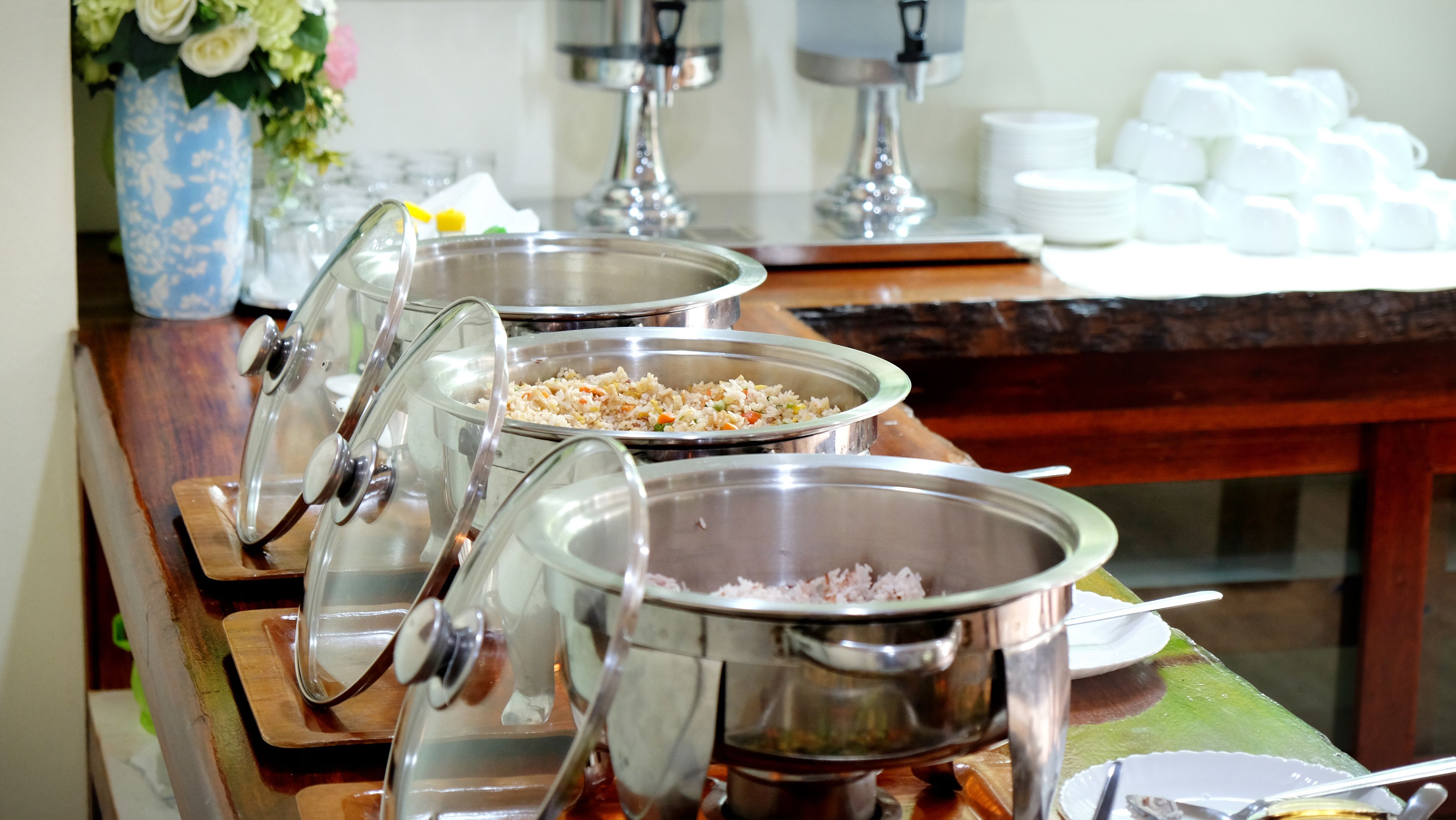 A buffet setup with multiple stainless steel chafing dishes containing food, a vase with flowers, and a stack of white plates and cups in the background.