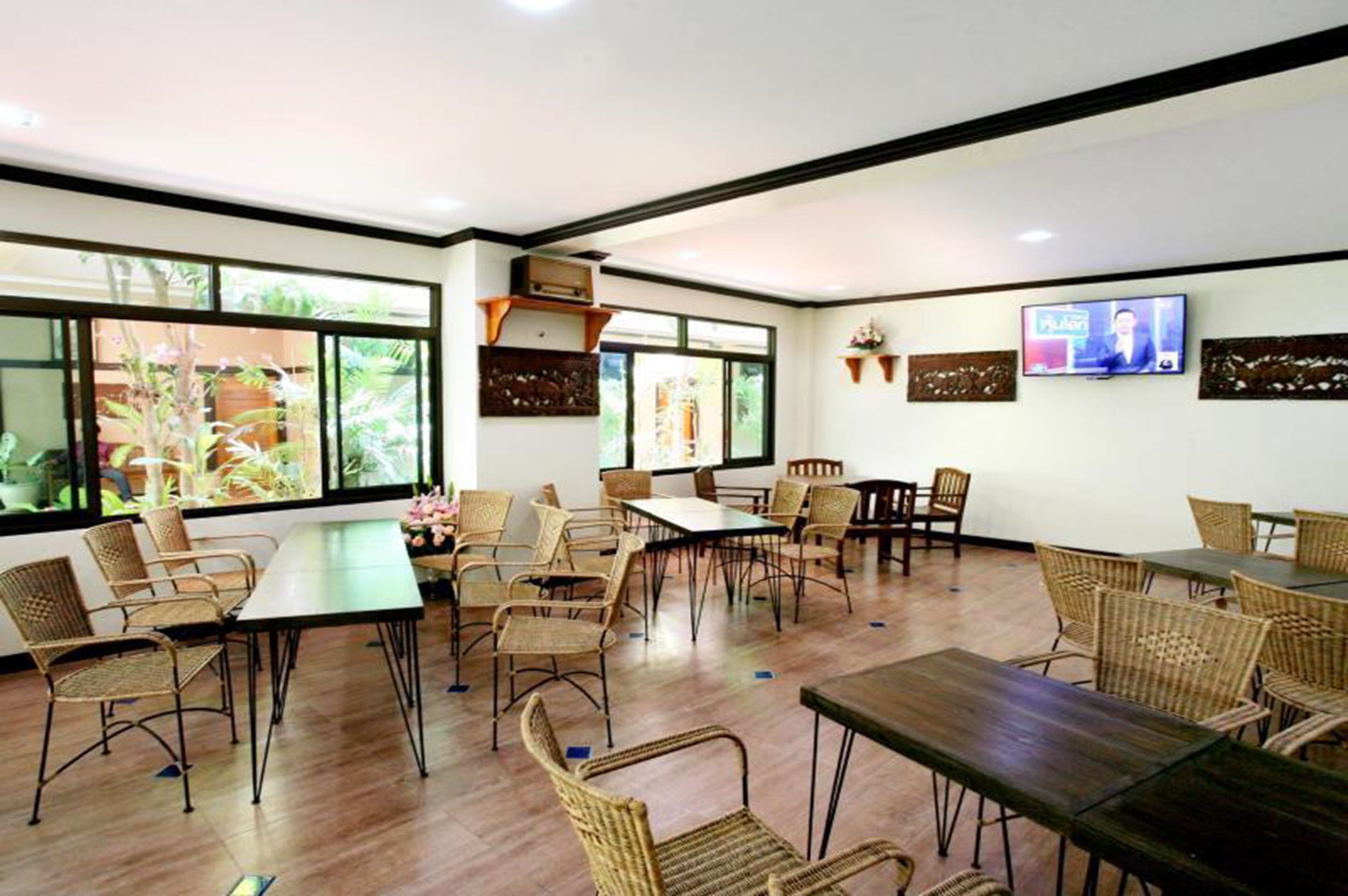 A spacious room with multiple tables and chairs, large windows with plants, and a television on the wall.