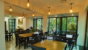 Uma área de refeições com várias mesas e cadeiras, grandes janelas com vista para o verde e luminárias pendentes em estilo lanterna.