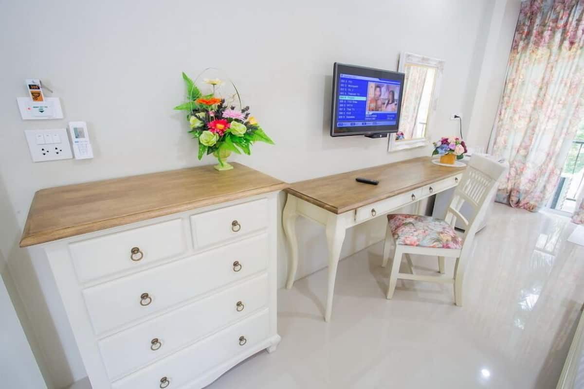 A room with a wooden desk, white drawers, a chair with floral upholstery, a television on the wall, and a floral arrangement on the desk.