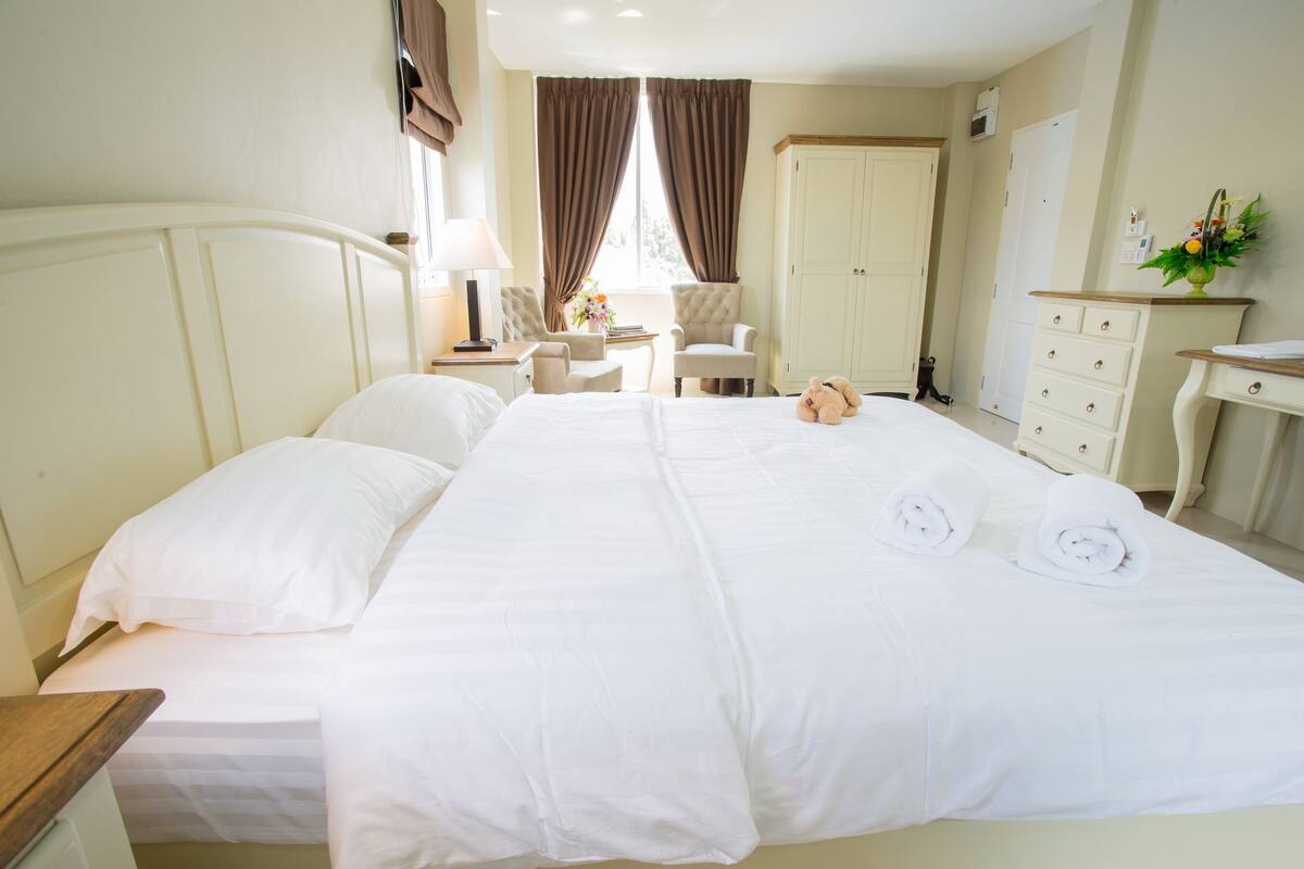 A neatly made bed with white linens, a bedside table with a lamp, a chair, and a wardrobe in a hotel room.