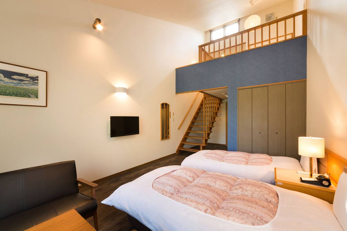 Ein Hotelzimmer mit zwei Betten, einer Couch, einem Fernseher, einer Wendeltreppe zu einer erhöhten Ebene und einem Gemälde an der Wand.