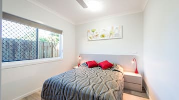 Un dormitorio con cama, mesita de noche, una obra de arte en la pared, ventilador de techo y ventana con vista a una valla.