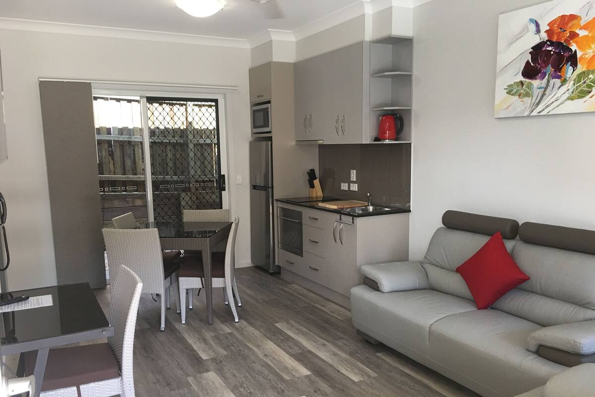 A modern living room with a grey sofa, a dining table with chairs, a kitchen area with a microwave, and a red kettle.