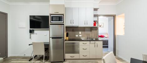 A modern kitchen with stainless steel appliances, a dining area with a wicker chair, and a view into a bedroom.