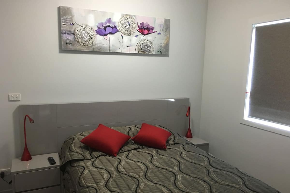A bedroom with a bed, two nightstands, a red lamp, a wall-mounted painting, and a window.