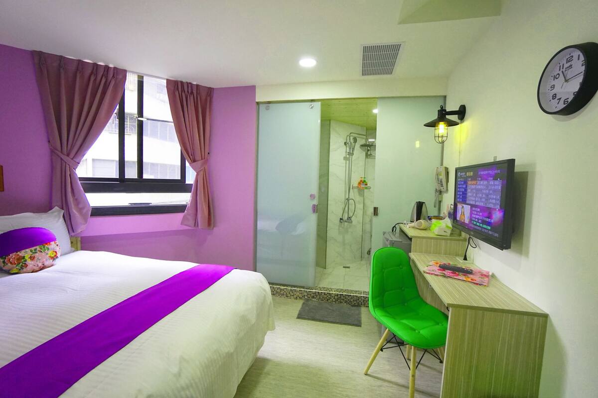 A hotel room with a bed, a desk with a computer, a green chair, a shower area, and a clock on the wall.