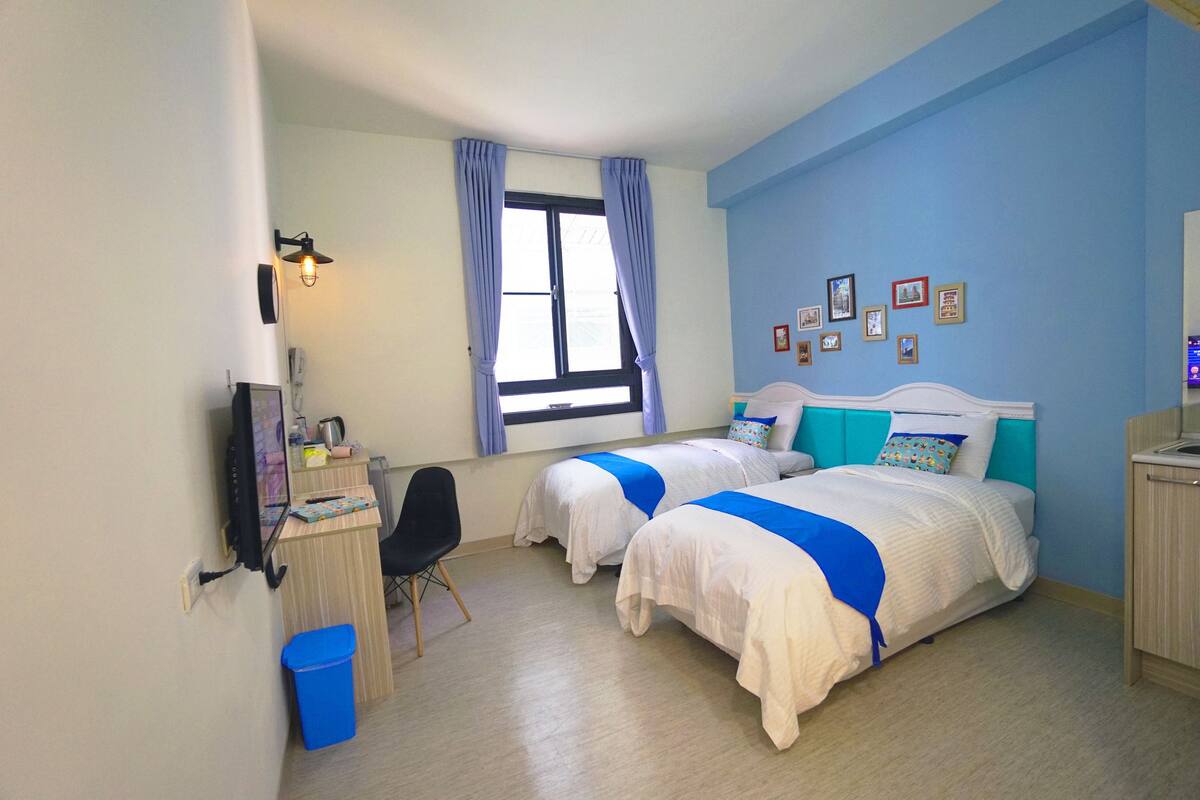 A bedroom with two beds, a desk, a chair, a TV, and a window with curtains.