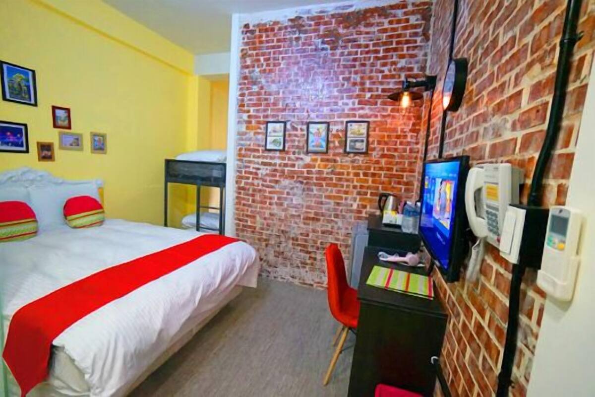 A hotel room with a brick wall, a bed with a red and white bedspread, a desk with a computer, and a red chair.