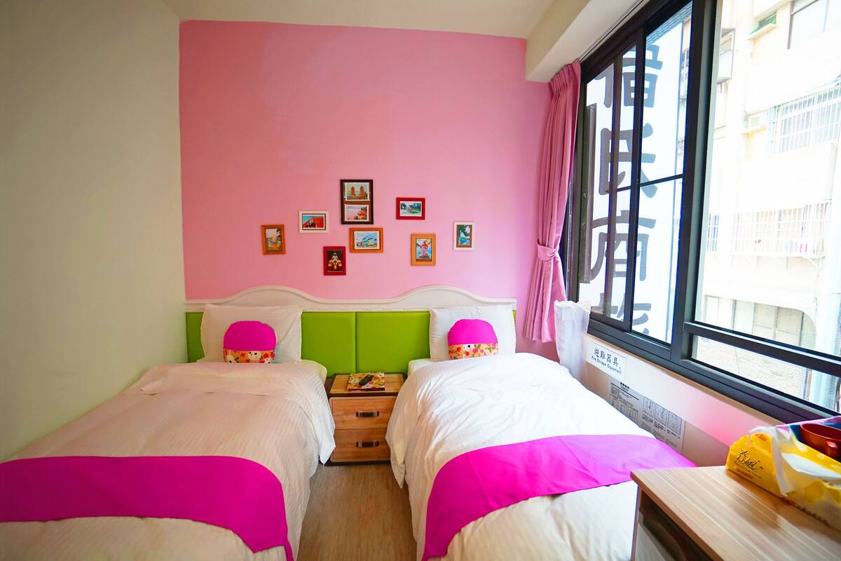 A room with two beds, a pink wall, and a window with a view of buildings.