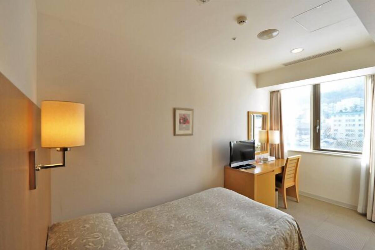 A hotel room with a bed, a desk with a computer, a lamp, and a window with a city view.