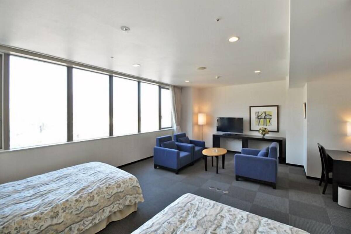 A hotel room with a bed, two blue armchairs, a small round table, a desk with a chair, a TV, and a large window.