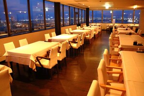 Una sala de conferencias con varias mesas y sillas, amplias ventanas que dan a una vista de la ciudad y suelo de madera.