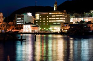 Un hotel frente al agua con varios pisos, un edificio destacado con ventanas iluminadas y un muelle con barcos.