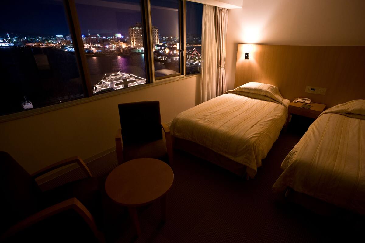 A hotel room with two beds, a chair, a small table, and a view of a cityscape at night.