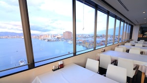 A restaurant with large windows offering a view of a harbor and cityscape. Tables are arranged for dining. - Winning Hotel (Hakodate)