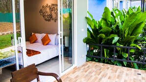 A bedroom with a bed, pillows, and a sliding glass door leading to a balcony with a view of a garden. - Chomphu Resort Khaolak (Takua Pa)