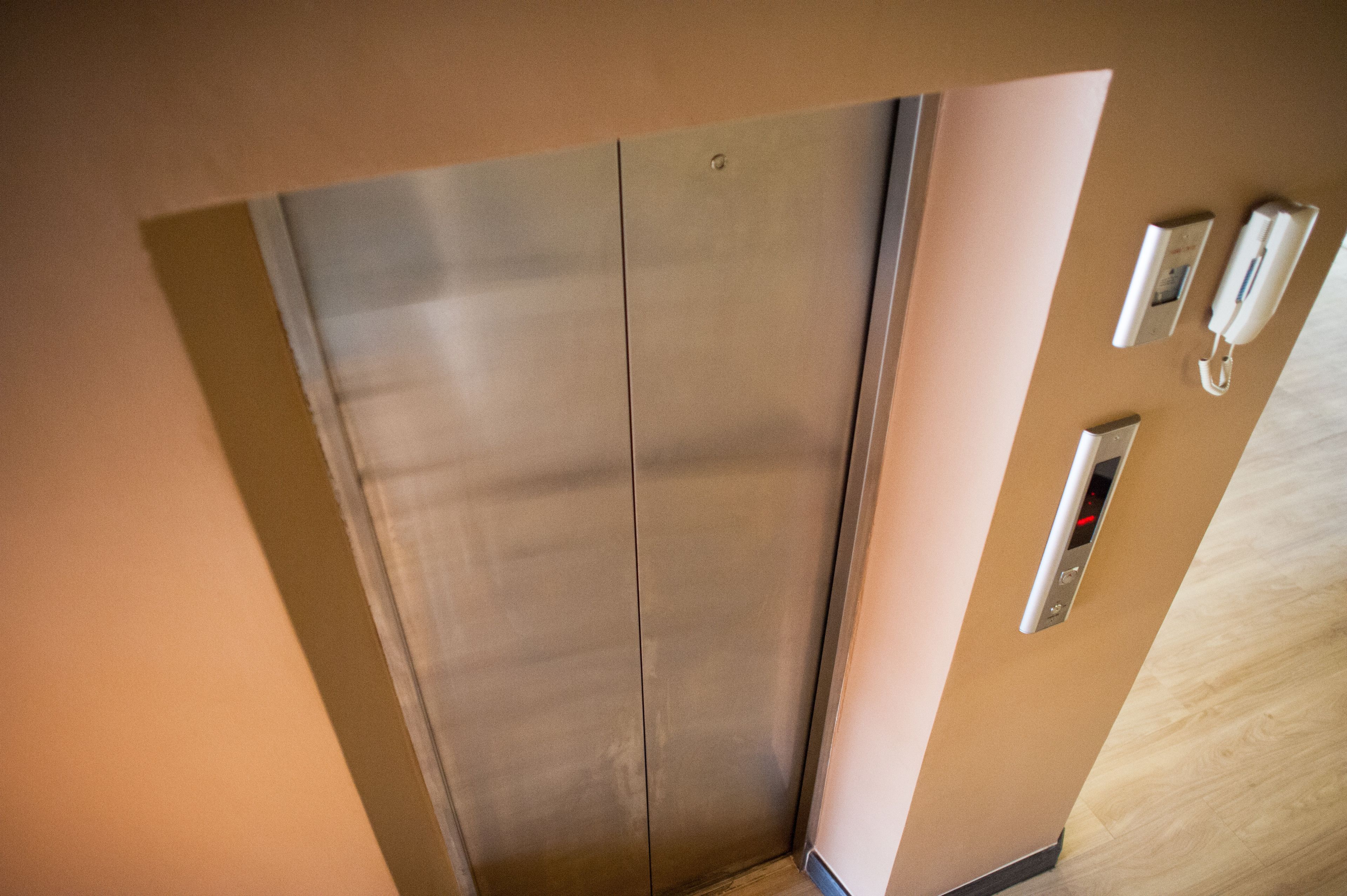 An elevator with a metallic finish, a wall-mounted intercom, and a control panel with a red light.