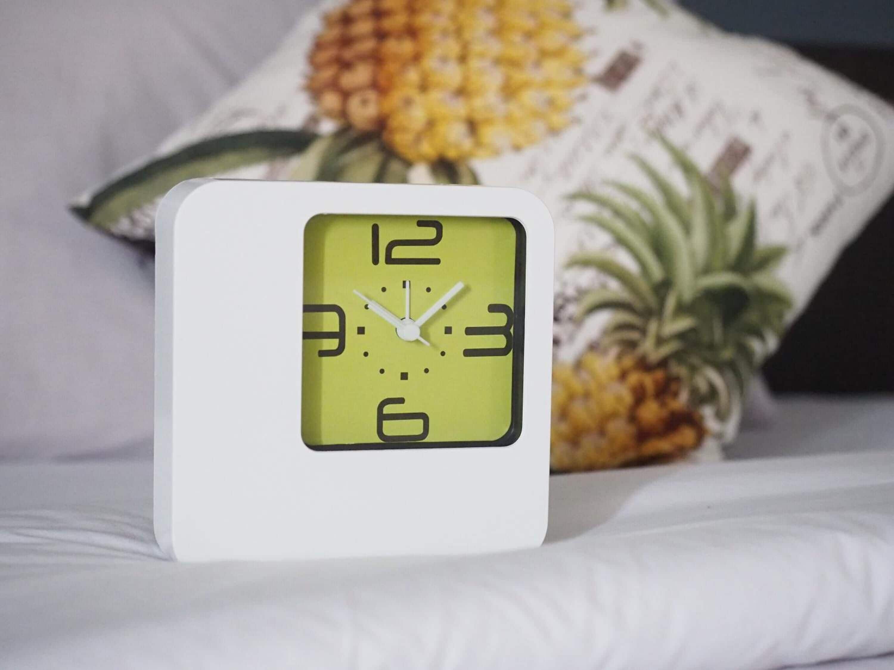 A white square alarm clock with a yellow face on a bed.