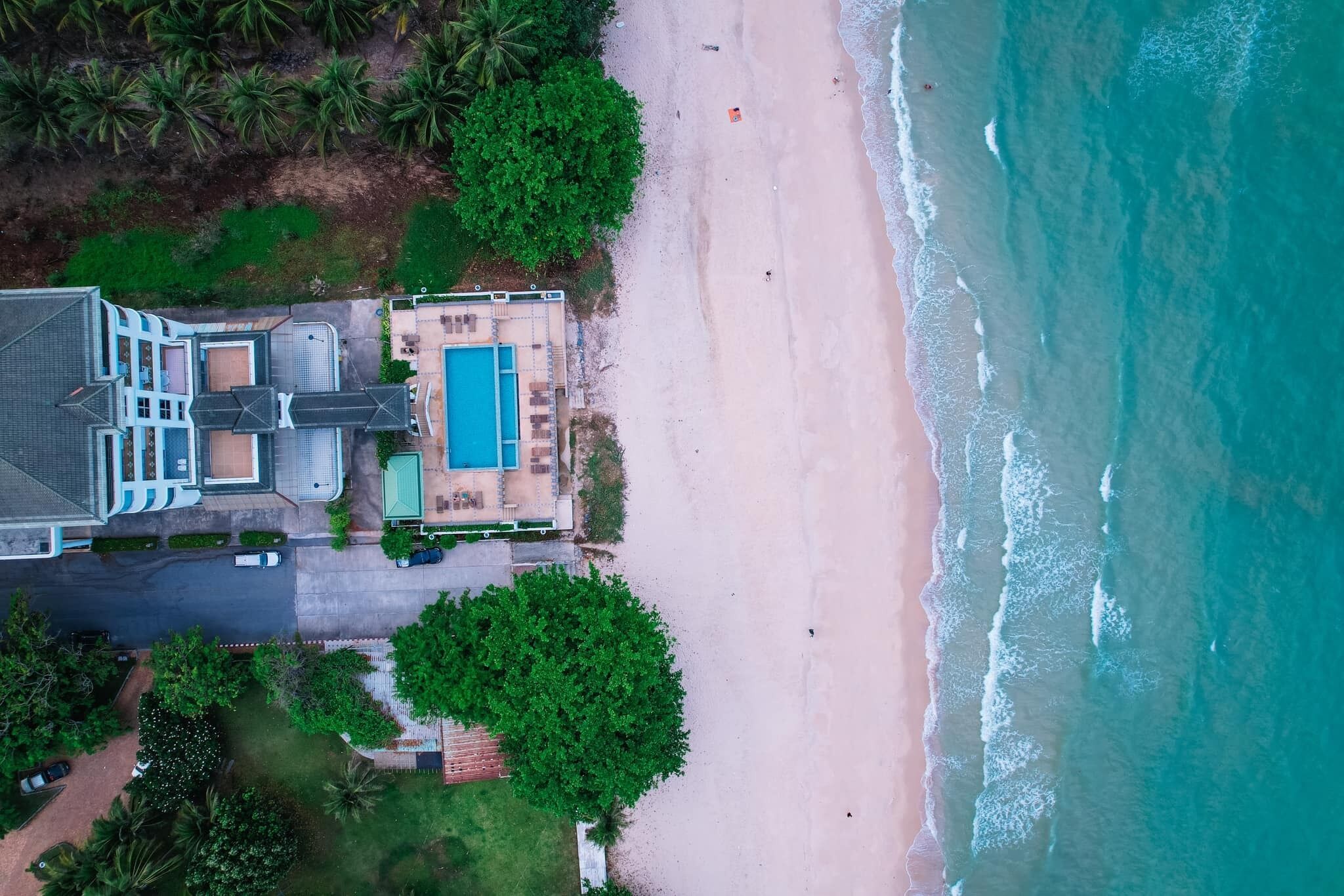 An aerial view of a beachfront property with a swimming pool, a large house, and a beach.