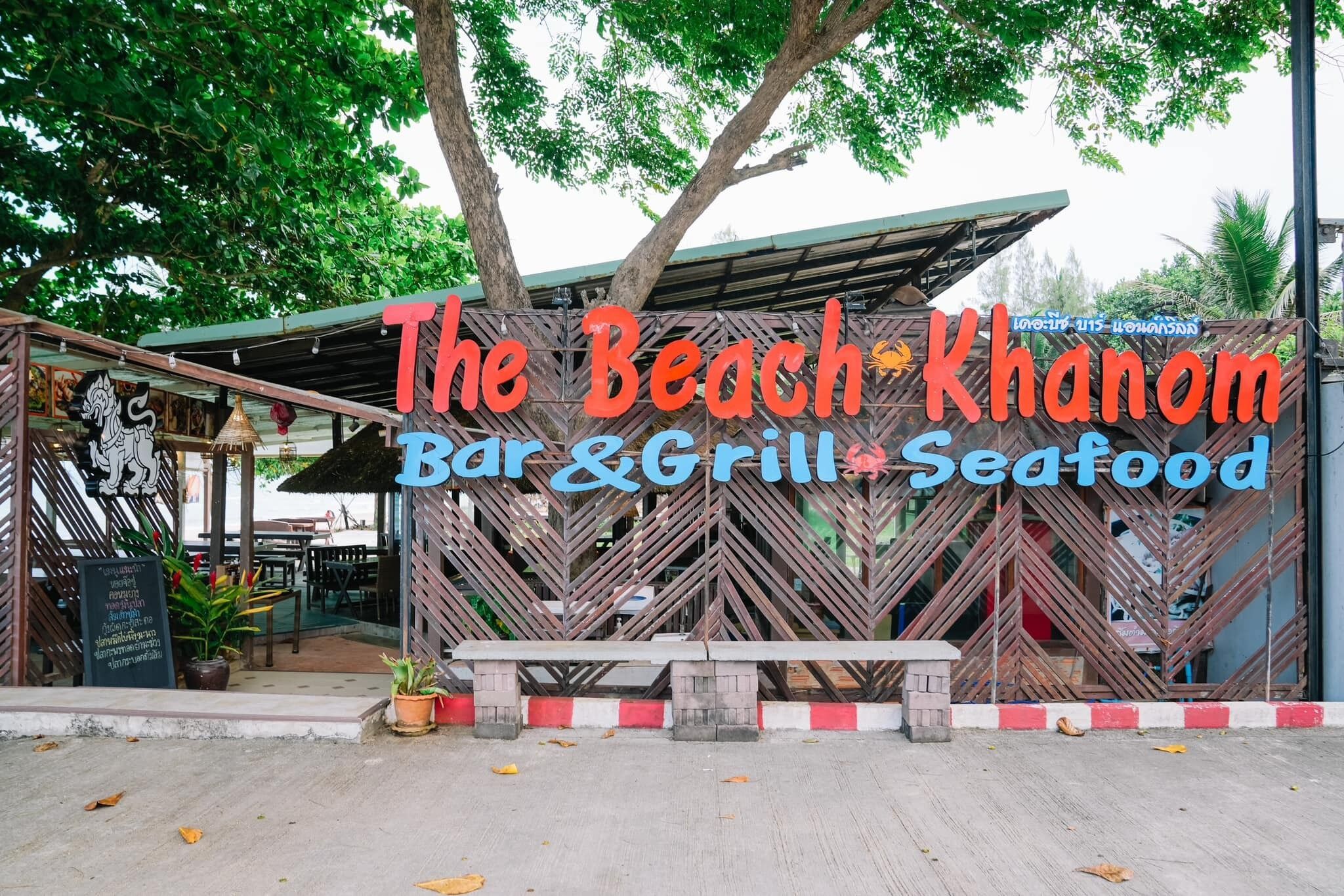 A beachside restaurant named "The Beach Khamon Bar & Grill" with a signboard and outdoor seating area.