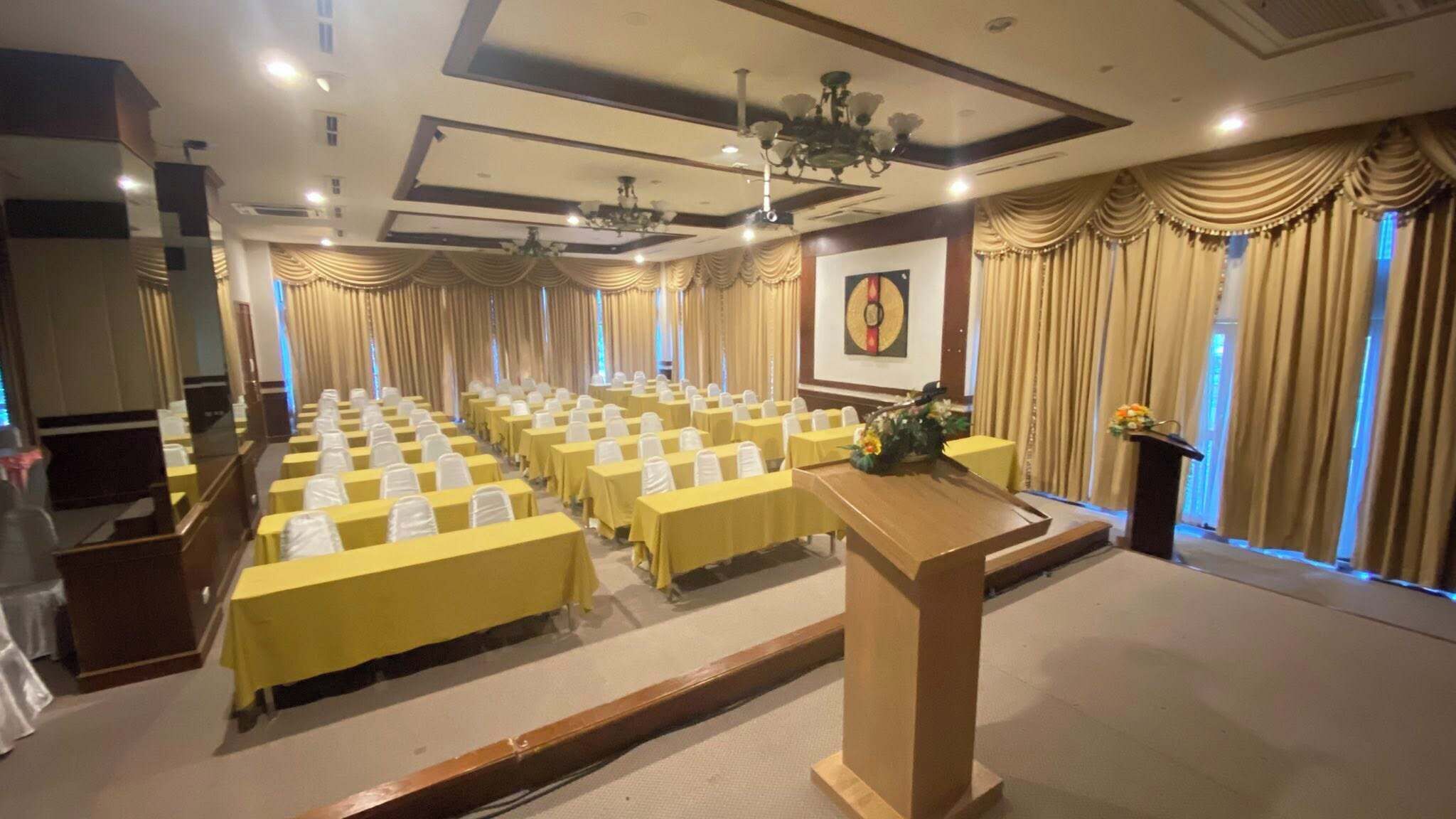 A conference room with rows of yellow tables and chairs, a podium, and a large window with curtains.