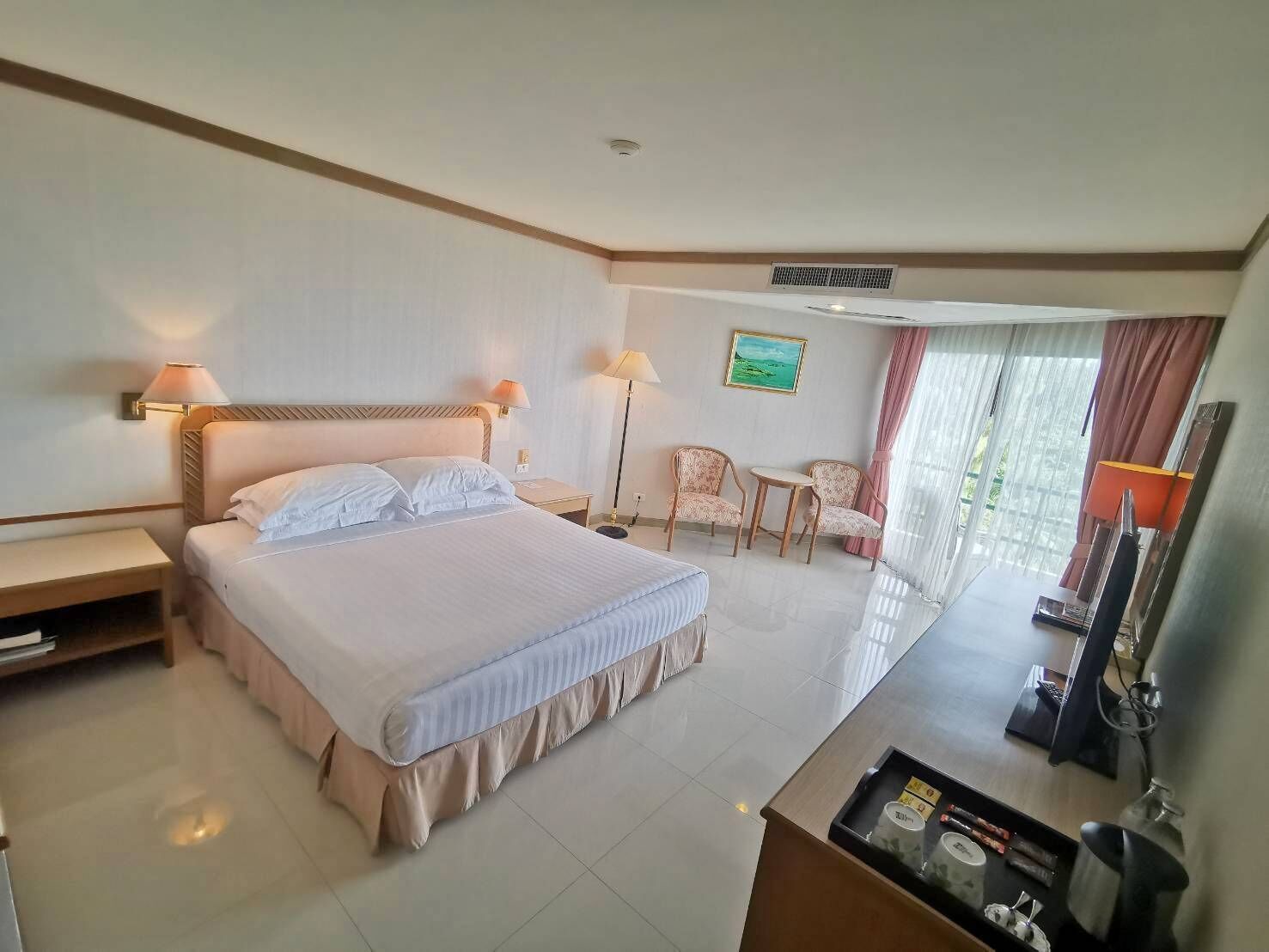 A hotel room with a large bed, two bedside tables, a desk with a computer, a chair, and a TV.