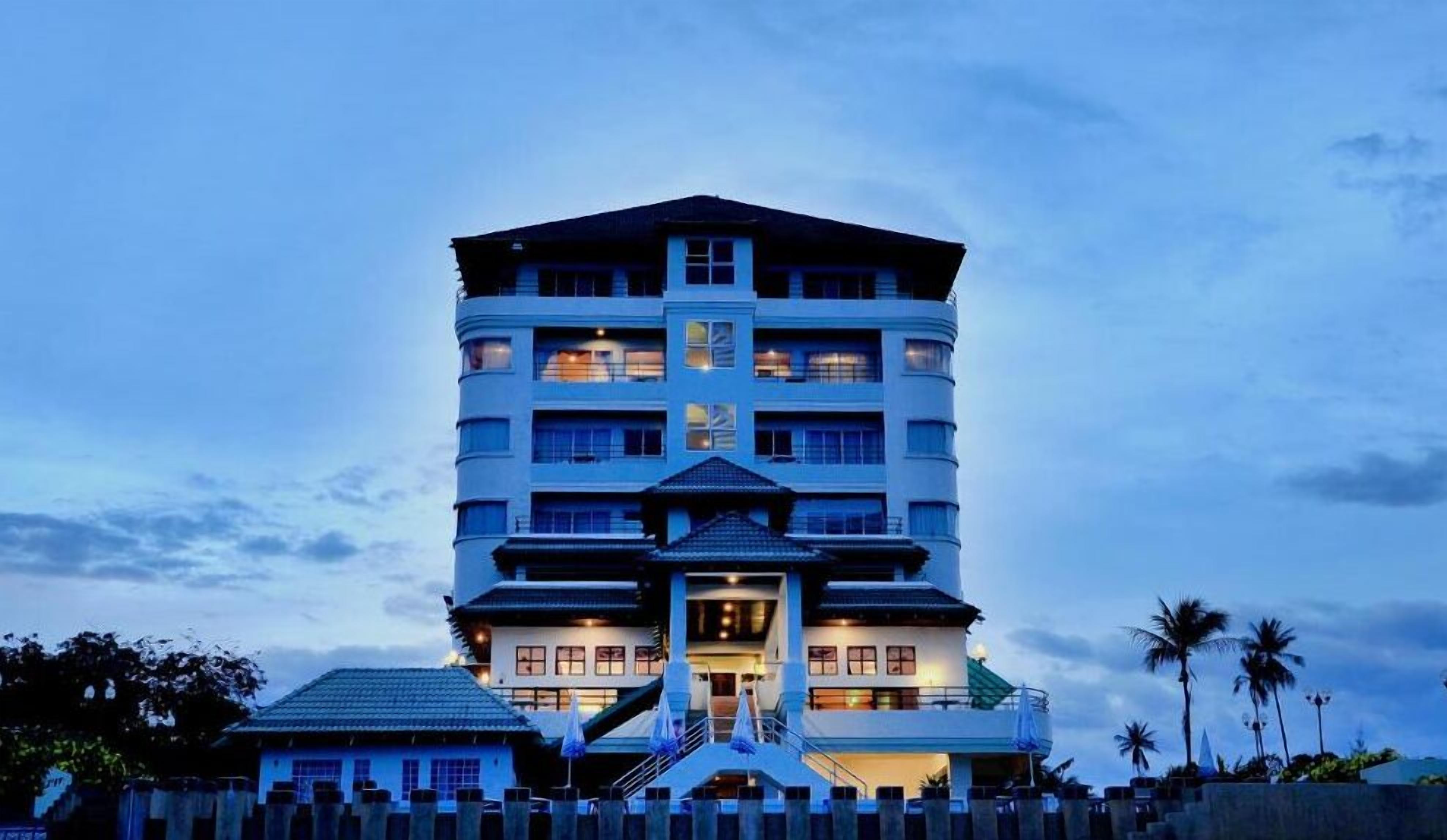 A multi-story hotel with a central entrance, balconies on each floor, and a surrounding fence.