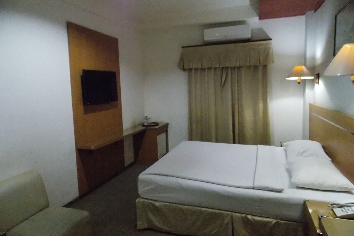 A hotel room with a bed, a desk, a chair, a TV mounted on the wall, and a window with curtains.