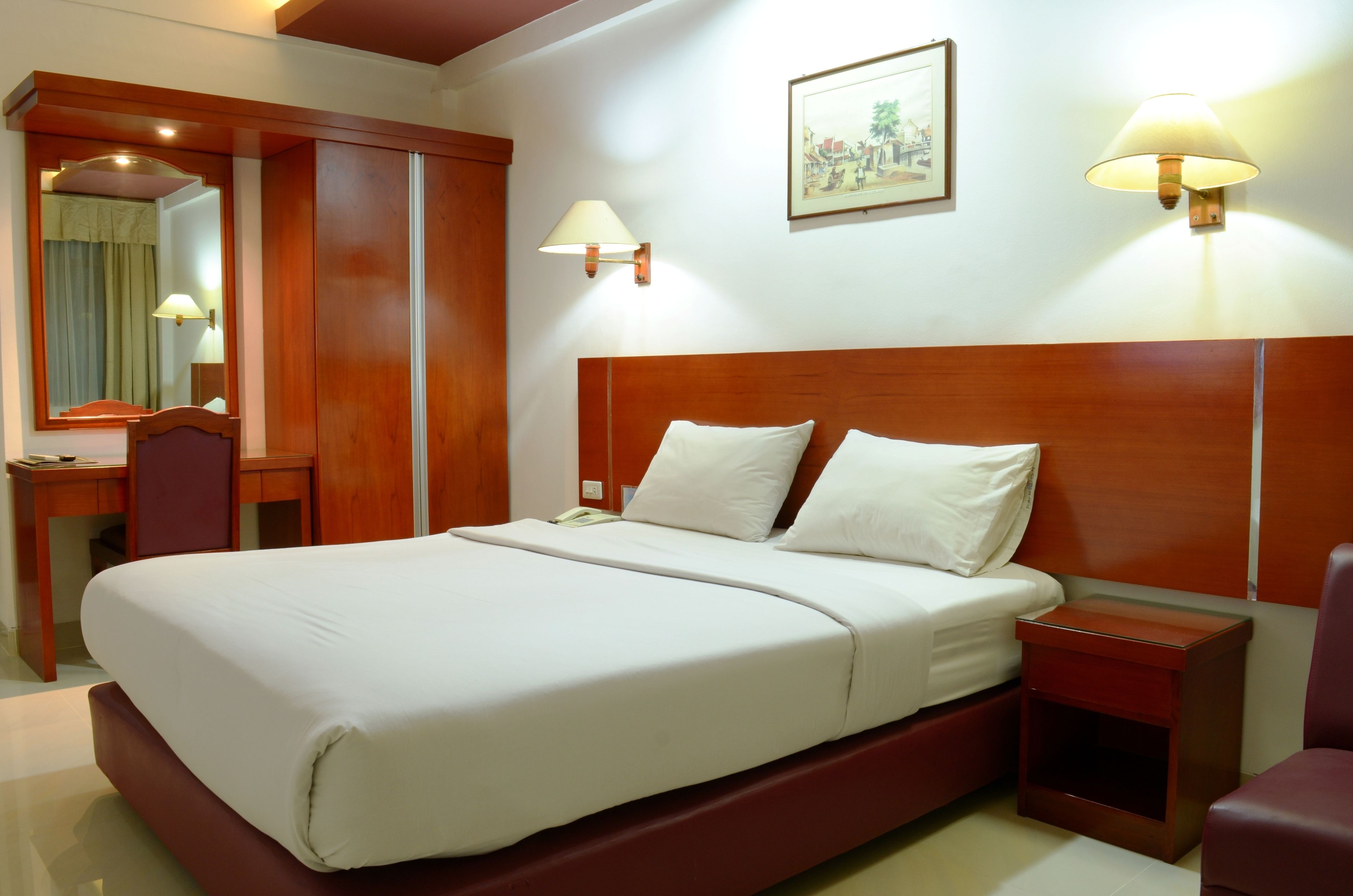 A hotel room with a wooden headboard, a large bed with white linens, a bedside table with a lamp, a mirror, and a framed picture on the wall.