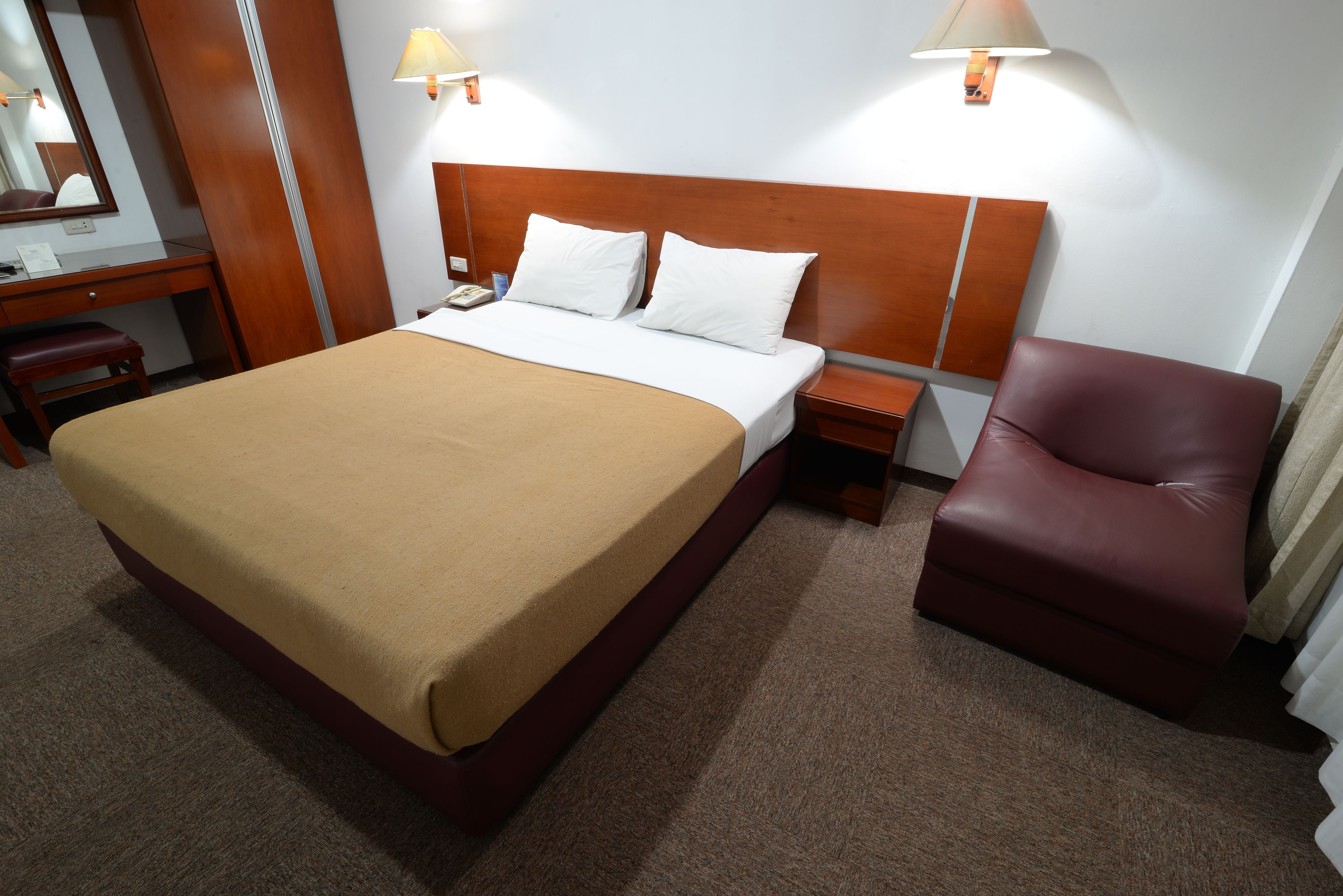 A hotel room with a large bed, a wooden headboard, two bedside lamps, a desk with a chair, and a leather armchair.
