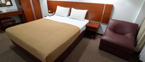 A hotel room with a large bed, a wooden headboard, two bedside lamps, a desk with a chair, and a leather armchair.